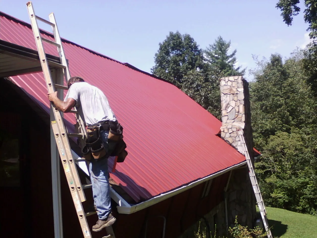 Roofer on a ladder beside a red metal roof and stone chimney — Soffit & Fascia Repair by Roofing Brothers in Sewaren